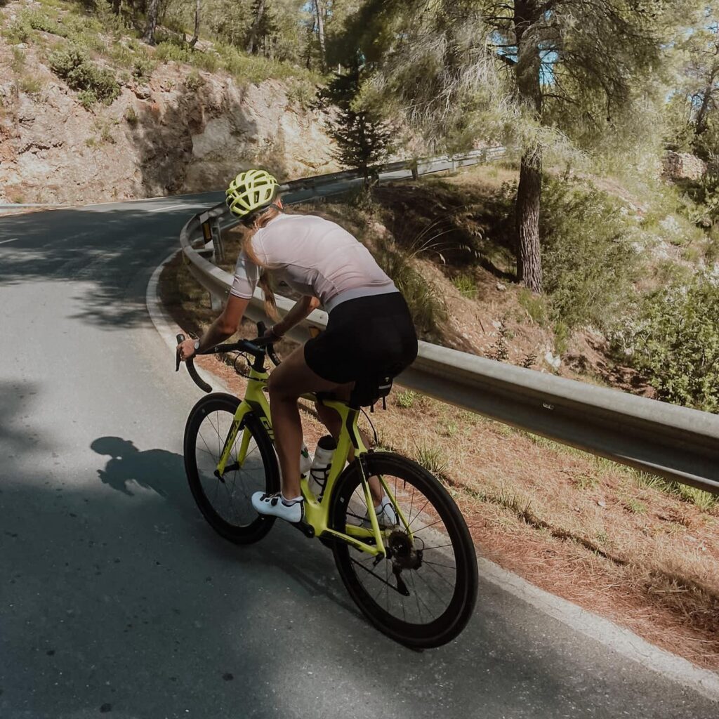 A cyclist in a yellow helmet and pink jersey is cruising on a yellow bike down a twisty road through the woods.