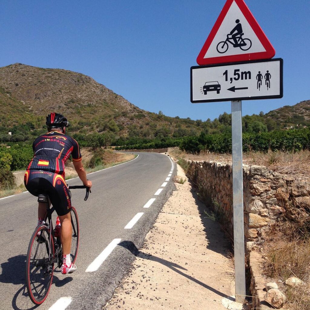 A cyclist is cruising down a country road with a sign reminding drivers to keep at least 1.5 meters away when passing bikes. The place is pretty scenic with hills and greenery all around.