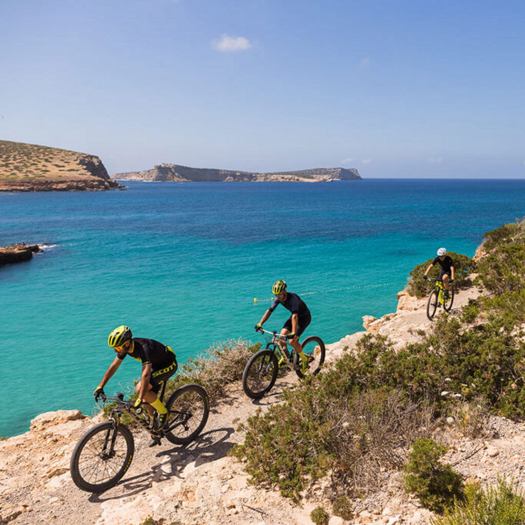 Three cyclists are cruising on a rocky coastal path, enjoying the clear blue sea and sunny skies.