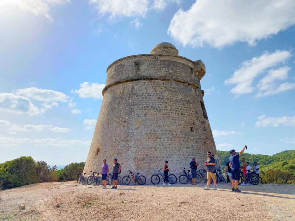 A bunch of bikers are hanging out with their bikes by this big round stone tower, and the sky’s all blue with some clouds around.