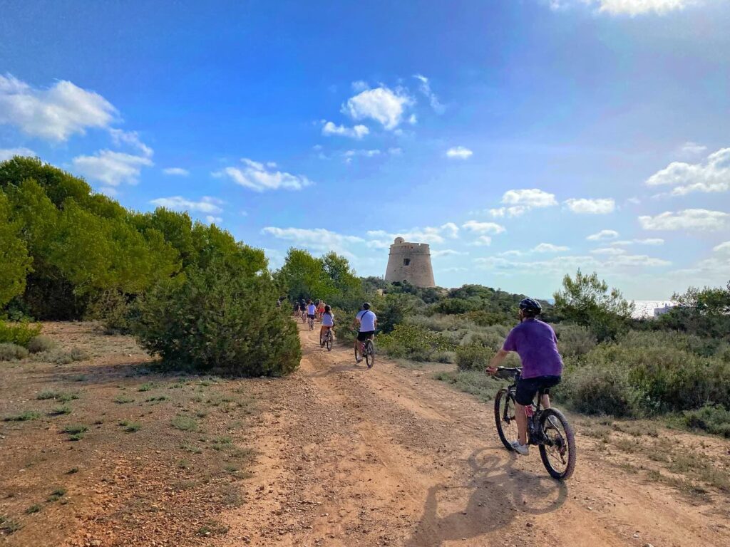 A bunch of folks are biking along a dirt trail, with lots of greenery and bushes around them. They're making their way to an old stone watchtower, all while the sky's partly cloudy.